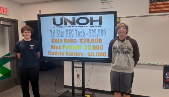 Two REC Tech students stand on either side of a white board with the names of scholarship winners listed. 